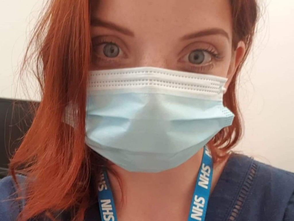 Woman with red hair wearing a mask and NHS lanyard taking a selfie