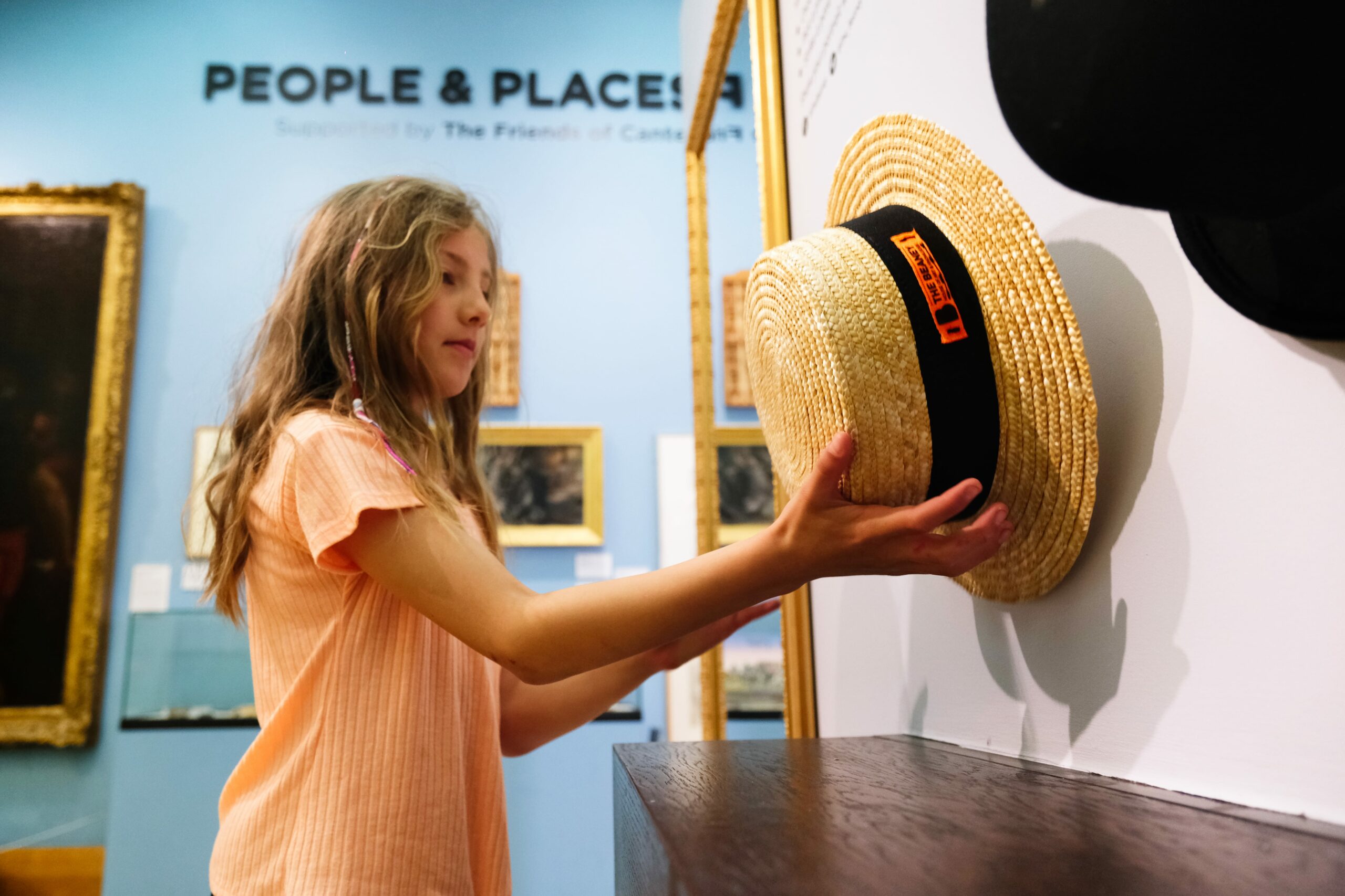 A young girl taking a straw boater hat from a hook in front of a mirror. On the blue gallery wall behind her are the words 'People & Places'