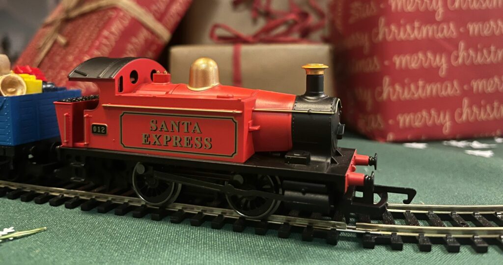 a red toy train with the words 'Santa Express' on the side in gold. It is running a long a track and in the background out of focus are presents wrapped in brow, gold and red paper.