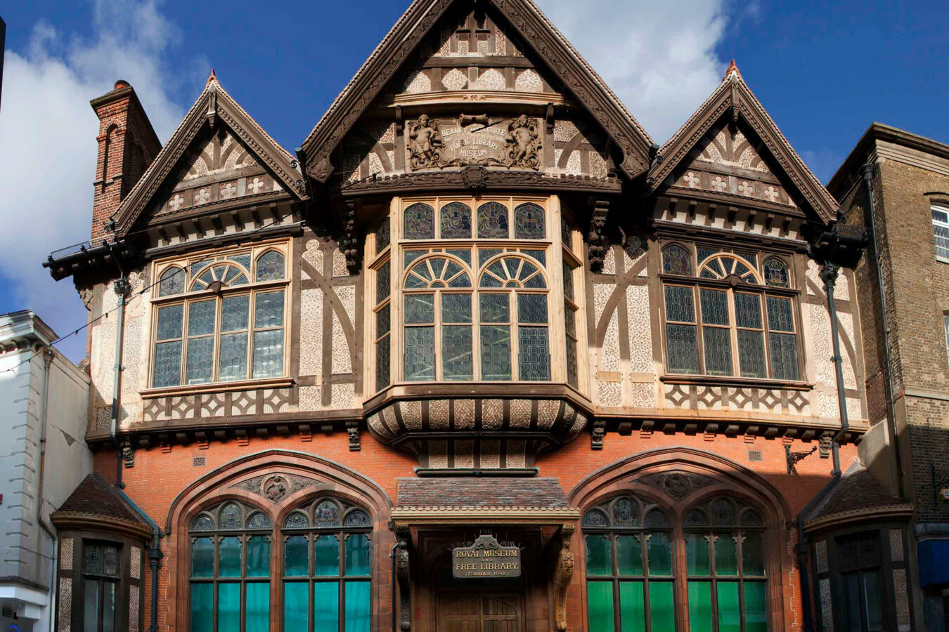 the facade of a grand tudor style building with large arched windows, decorative woodwork and three triangular roofs.