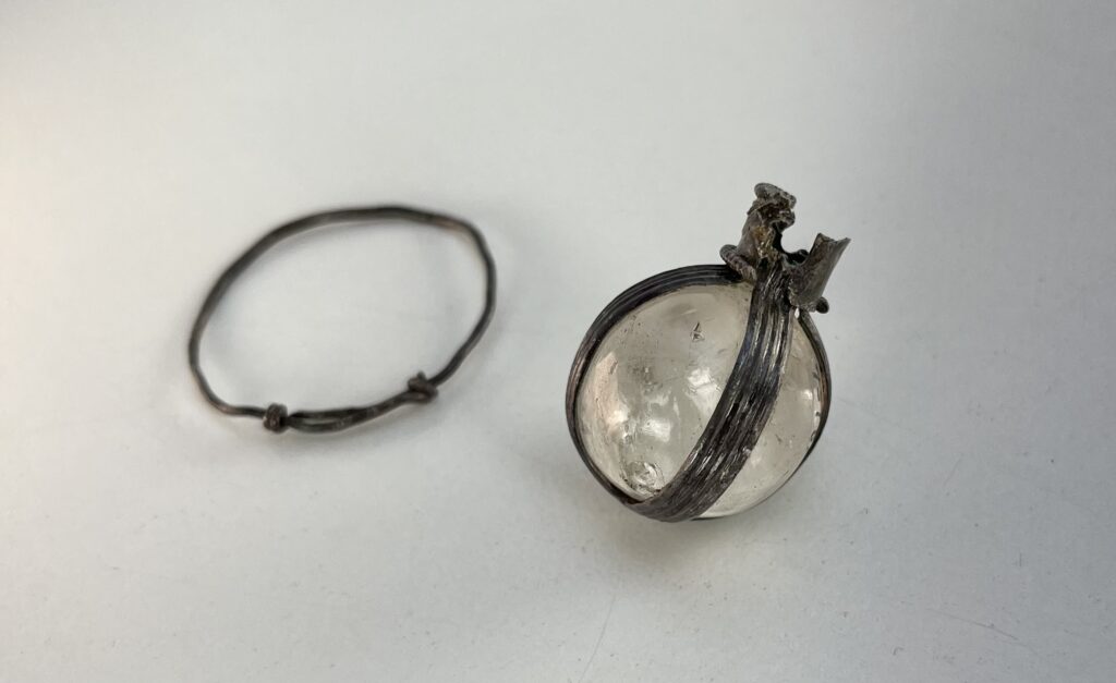 A crystal spehere surrounded by an aged metal sling with grooves. Next to it on the left is a metal loop that has broken from the object