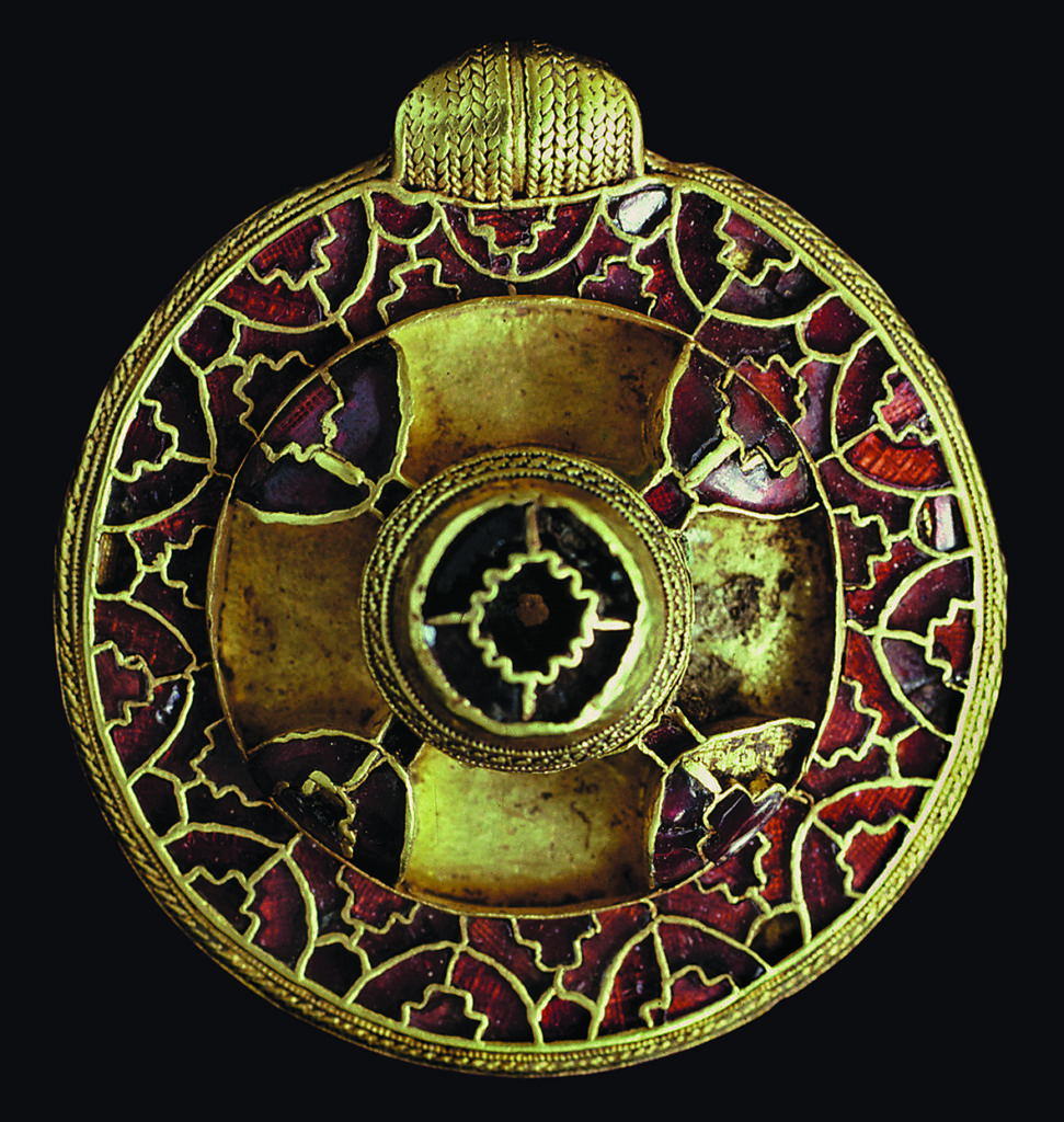 a round golden metal broch with intricate red cut stone inlaid in patterns and a central circular design