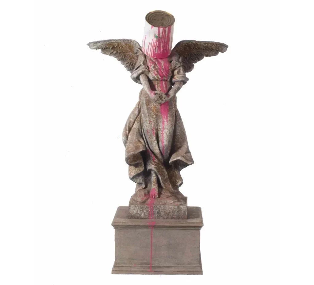 Statue of an angel with wings, holding their hands in front of them. There is an upside paintpot on their head spilling pink paint over the statue